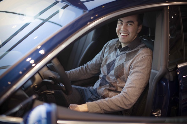En mann som sitter i førersetet av en bil, iført en grå argyle-genser og smiler til kameraet.